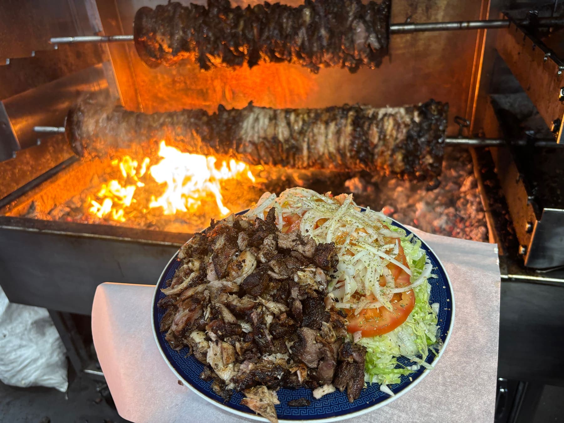 A plate of charcoal meat and salad in front of the spits turning over open flame